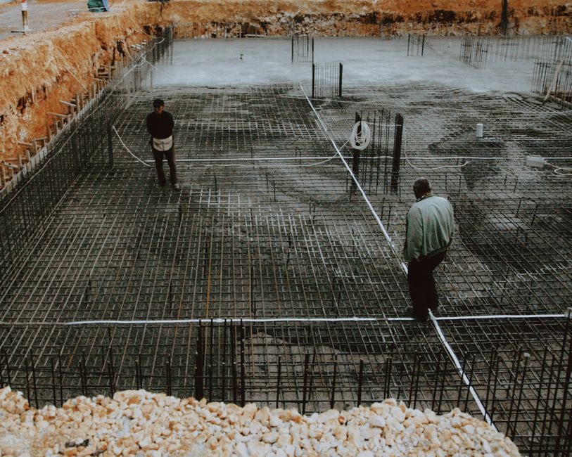 Two workers inspecting a rebar grid foundation at a construction site.